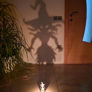 Puede incluir: Una figurita de bruja blanca proyecta una sombra en una pared, creando una escena de Halloween espeluznante. La figurita está iluminada por una fuente de luz, creando un brillo cálido.