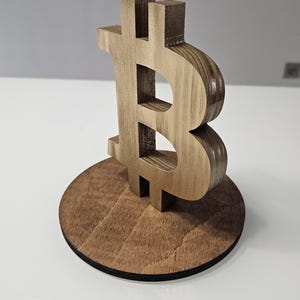 Puede incluir: Un símbolo de Bitcoin de madera sobre una base redonda de madera. El símbolo es marrón y tiene una superficie texturizada.