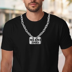 May include: Black t-shirt with a silver chain necklace and a rectangular pendant. The pendant has the text "MIAMI 305" in a bold, sans-serif font. The background shows a street scene with buildings and palm trees.
