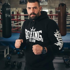 Puede incluir: Un hombre con una sudadera negra con "BOXING CLUB" impreso en la parte delantera y la manga, de pie en un gimnasio de boxeo. El gimnasio tiene sacos de boxeo, un ring y carteles en la pared. Lleva un reloj y tiene los puños cerrados.
