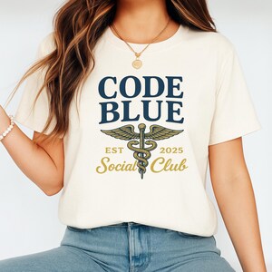 May include: Cream-colored t-shirt with the text "CODE BLUE" in navy blue, above a caduceus symbol with wings. Below the symbol are the words "EST 2025 Social Club" in gold. The shirt is worn by a person in blue jeans.
