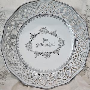 May include: White ceramic plate with a silver filigree design around the edge. The center of the plate has a silver inscription that reads "Zur Silberhochzeit."