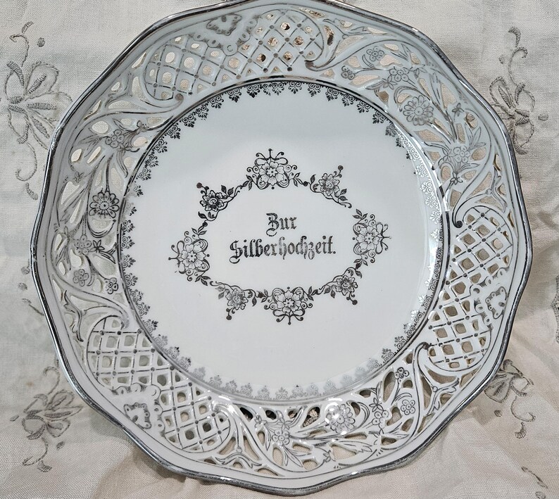 May include: White ceramic plate with a silver filigree design around the edge. The center of the plate has a silver inscription that reads "Zur Silberhochzeit."