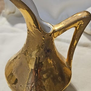 May include: A small, gold-colored pitcher with a textured surface and a white interior. The pitcher has a curved spout and a handle. The gold finish gives the pitcher a vintage or antique appearance. The pitcher is likely made of ceramic or porcelain.