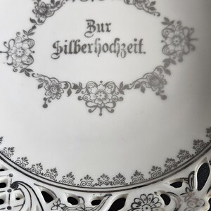 May include: A white ceramic plate with a silver design. The plate has a scalloped edge and a cut-out design in the center. The words "Zur Silberhochzeit" are written in silver on the plate.