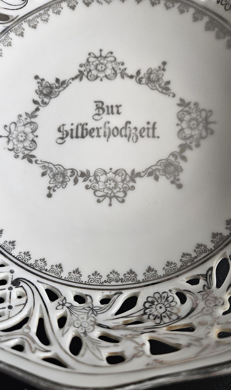 May include: A white ceramic plate with a silver design. The plate has a scalloped edge and a cut-out design in the center. The words "Zur Silberhochzeit" are written in silver on the plate.