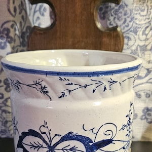 May include: A white ceramic container with blue floral designs and a wooden wall mount. The container has a cylindrical shape with a flared base and rim. The blue designs include leaves and ribbons.