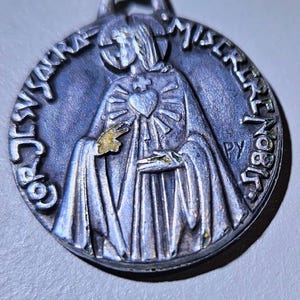 May include: A circular silver-toned metal pendant featuring a relief of a figure with a heart on the chest. The text COR JESU SACRA MISERERE NOBIS is embossed around the edge, with the letters PY visible on the right side of the figure.