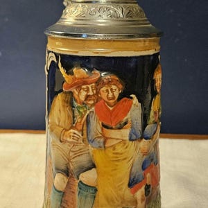 May include: A ceramic beer stein with a silver-colored metal lid. The stein features a detailed relief scene of people in traditional clothing. The colors include blue, yellow, brown, and green.
