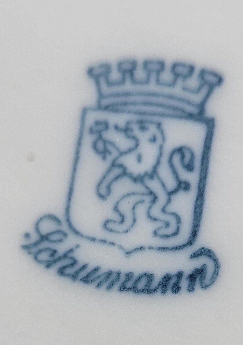 May include: A blue and white porcelain mark with a crowned lion and the text "Schumann" underneath.