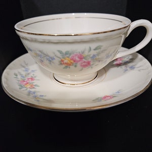 May include: A white teacup and saucer set with a floral pattern in shades of pink, yellow, and blue. The set has a gold trim around the edges.