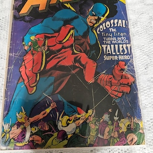 May include: A comic book cover featuring a superhero named Atom, who is depicted as a giant, with a red and blue costume, holding a group of smaller figures in his hand. The cover text reads "Colossal! The Tiny Titan Turns Into The World's Tallest Super-Hero!"