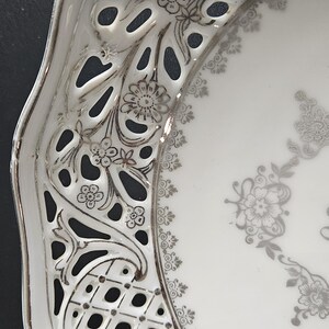 May include: A white porcelain plate with a silver filigree design. The plate has a scalloped edge and a delicate floral pattern.