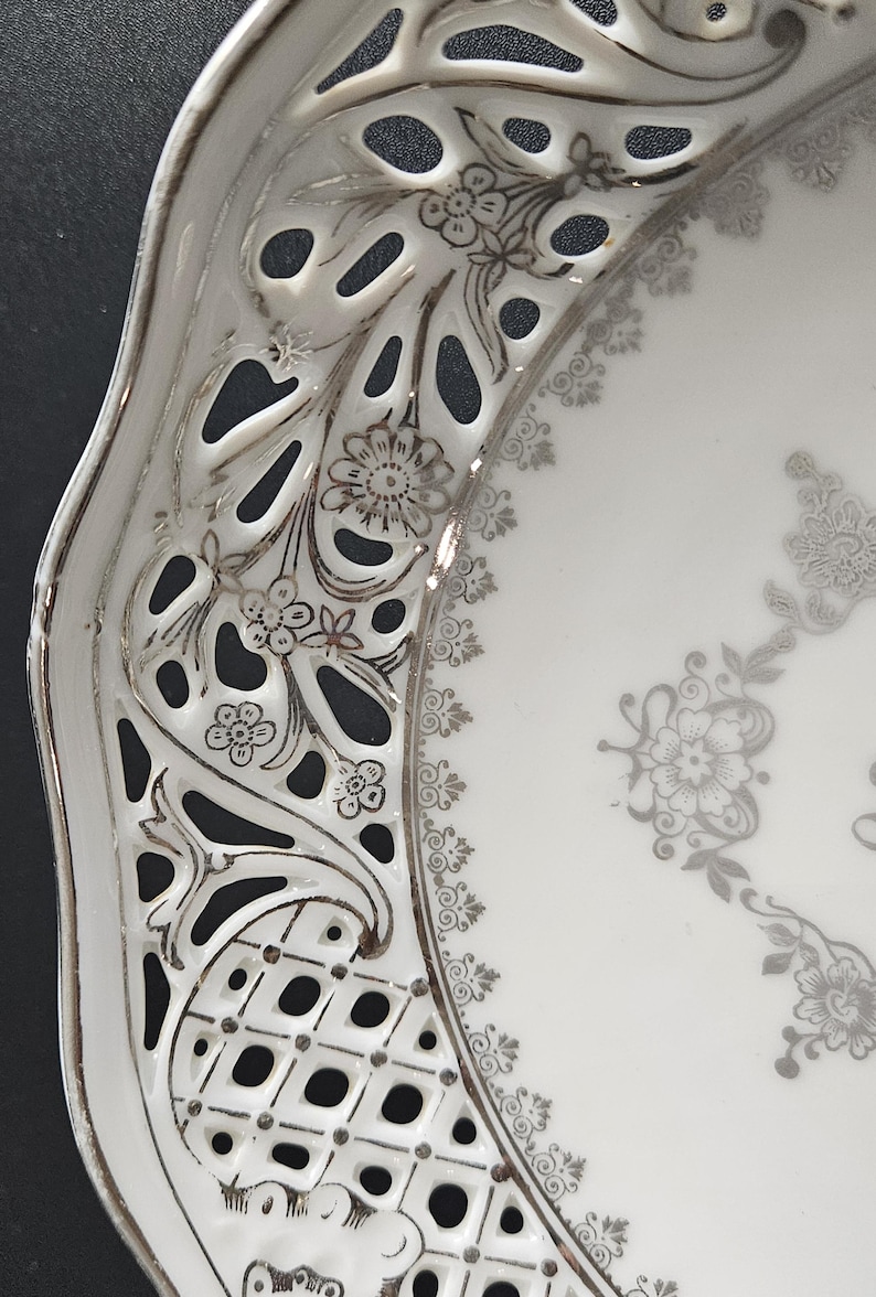May include: A white porcelain plate with a silver filigree design. The plate has a scalloped edge and a delicate floral pattern.