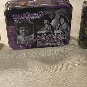 May include: Three vintage-style metal lunch boxes. Each box features a different image of children and the text "Our Gang". The boxes have a rectangular shape with a handle and a clasp closure.