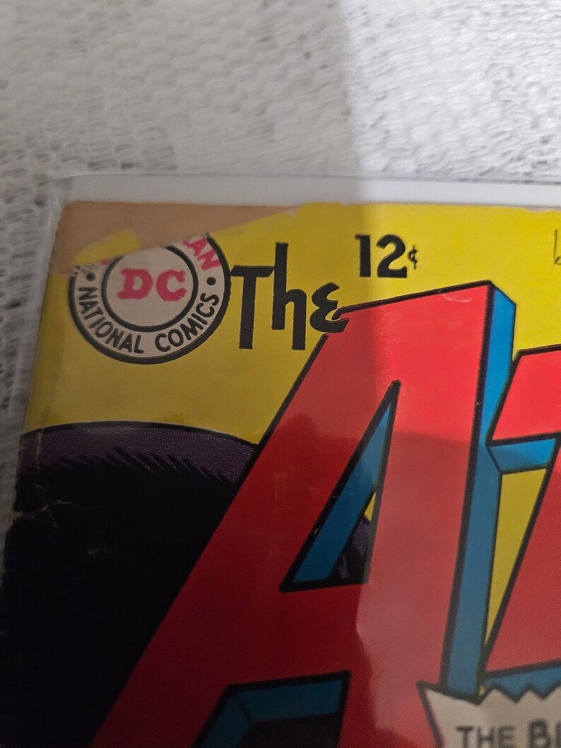 K&ouml;nnte beinhalten: Ein Vintage-Comic-Cover mit einem roten, blauen und gelben "A" mit dem Text "The" und "12&cent;" dar&uuml;ber. Das Cover zeigt auch einen runden Aufkleber mit dem Text "National Comics" und "DC" in einem roten Kreis.