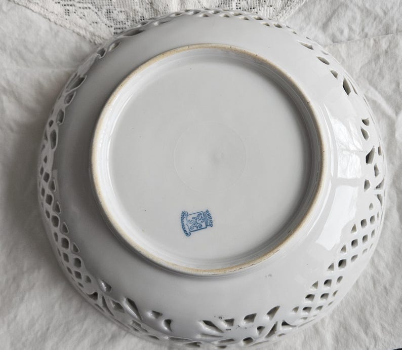 May include: White ceramic bowl with a delicate, openwork design. The bowl has a round shape and a smooth, white glaze. The bottom of the bowl features a blue and white logo with the text "Germany" in blue.