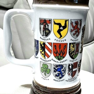 May include: A white ceramic beer stein with a brown rim and base, featuring colorful crests of various German cities. The stein has a large white handle and is likely used for drinking beer. The design includes city names like Bamberg and Passau.