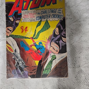 May include: A comic book cover featuring a superhero called The Atom. The cover is yellow with green and red accents. The Atom is fighting a villain wearing a purple mask. The text on the cover reads "The Challenge of the Computer Crooks!"