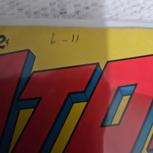 K&ouml;nnte beinhalten: Ein rotes und gelbes Comic-Cover mit dem Wort "ATOM" in gro&szlig;en, fetten, blau umrandeten Buchstaben. Das Cover hat eine handschriftliche Nummer "6-11" in der oberen rechten Ecke.