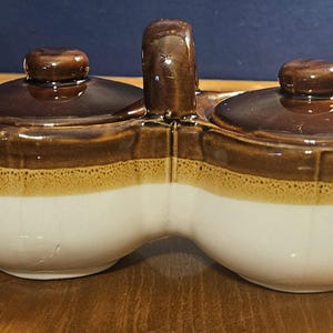 May include: A ceramic salt and pepper shaker set with two connected compartments and a handle. The set features a white base, a tan band, and a dark brown lid and handle. The design is a classic, vintage style.