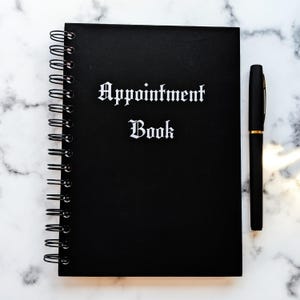 Puede incluir: Un libro de citas negro con espiral con las palabras "Appointment Book" en letras góticas blancas. Un bolígrafo negro con detalles dorados descansa junto al libro sobre una superficie con patrón de mármol. El libro está cerrado.