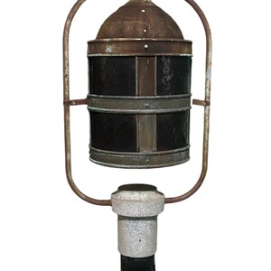 May include: A vintage, rusty metal street lamp with a black and brown lantern. The lantern has a dome-shaped top and is suspended from a curved metal frame. The lamp post is made of concrete and has a black base.