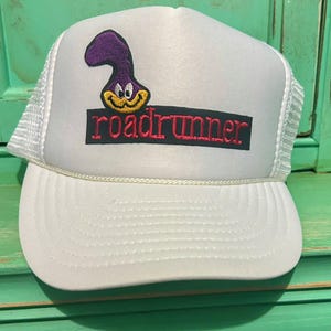 Può includere: Cappellino da camionista bianco con una toppa ricamata nera e rossa che dice "roadrunner". La toppa presenta anche un roadrunner dei cartoni animati con la testa viola e il becco giallo.