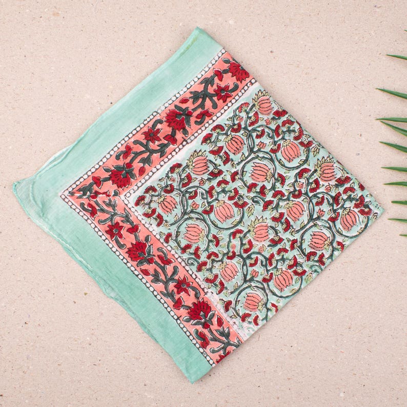 May include: A square, folded textile with a floral pattern. The border is a light teal color, with a coral and red floral design. The center features a repeating pattern of coral flowers and green vines on a light teal background. The textile is likely cotton.