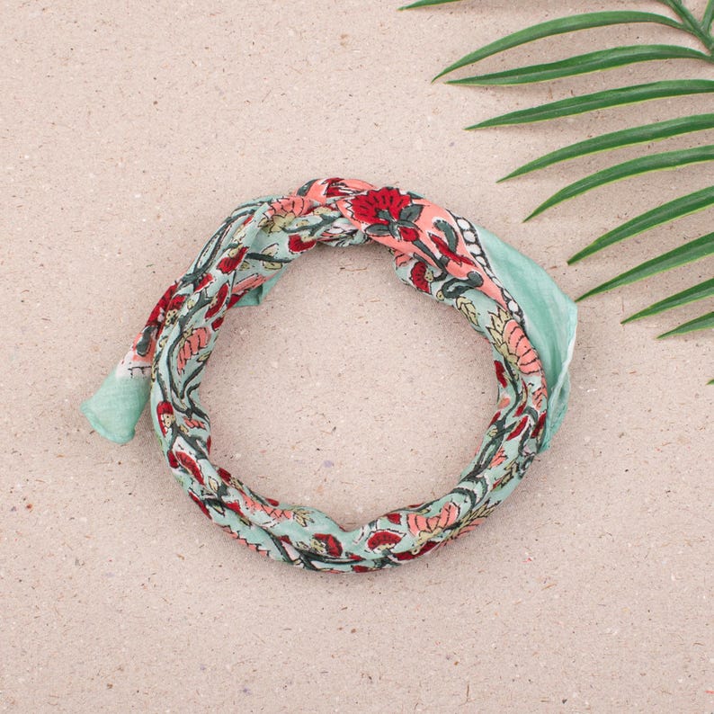 May include: A floral patterned headband in shades of red, pink, and green. The headband is made of a soft fabric and is tied in a knot. The background is a light tan color with a green leaf in the upper right corner.