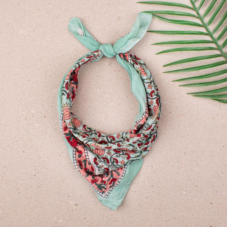 May include: A square bandana with a floral pattern in red, pink, and green on a light blue background. The bandana is tied at the top, showcasing the intricate design. The fabric appears soft and lightweight.