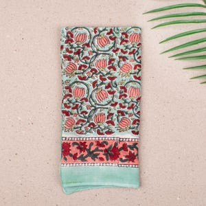 May include: A folded, rectangular cloth with a floral pattern. The main design features pink flowers and green leaves on a light blue background. The bottom border has a coral band with red floral accents, and a light green edge.
