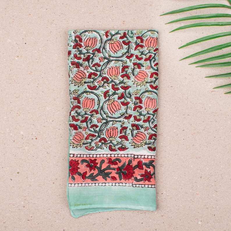 May include: A folded, rectangular cloth with a floral pattern. The main design features pink flowers and green leaves on a light blue background. The bottom border has a coral band with red floral accents, and a light green edge.