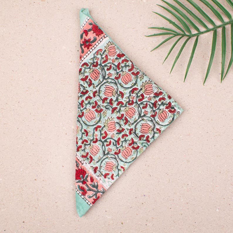 May include: A folded, triangular cloth napkin with a floral pattern. The napkin features a light blue background with red and pink floral designs and a red and pink border. The napkin is made of a soft fabric.