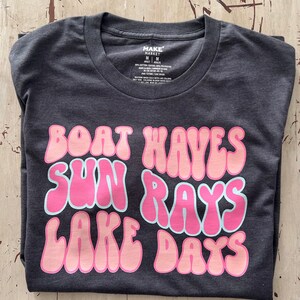 Peut inclure: Un t-shirt gris fonc&eacute; avec un motif rose et bleu clair qui dit "Boat Waves Sun Rays Lake Days".
