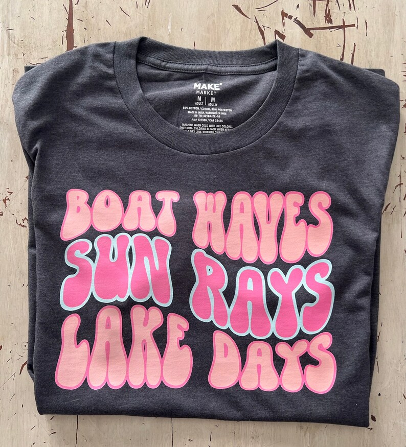 Peut inclure: Un t-shirt gris fonc&eacute; avec un motif rose et bleu clair qui dit "Boat Waves Sun Rays Lake Days".