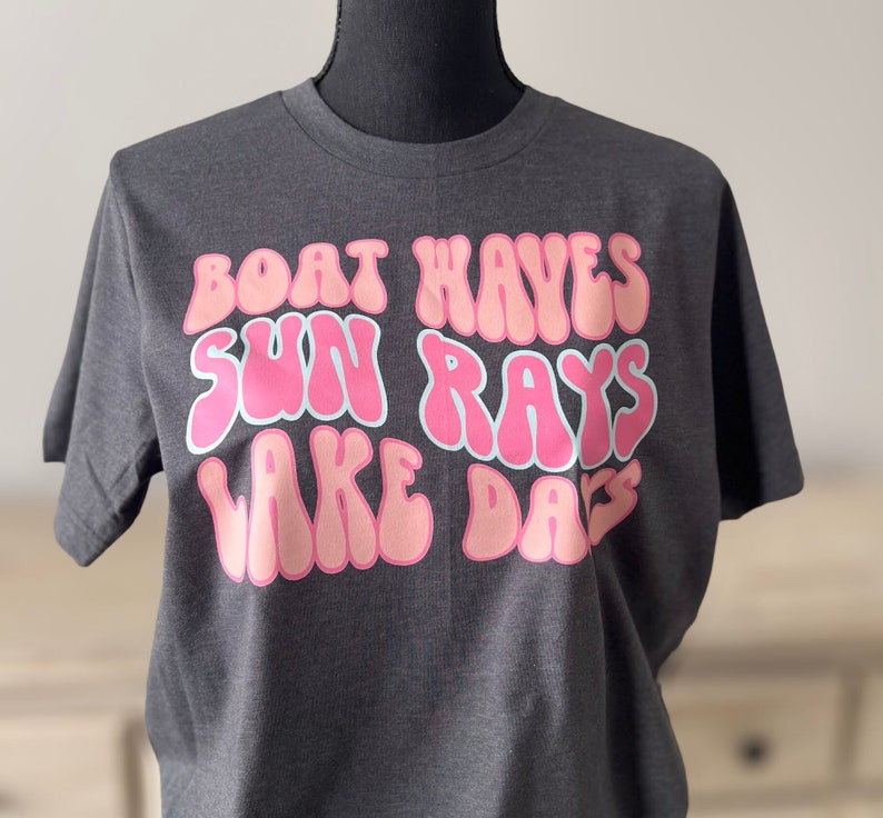 Peut inclure: Un t-shirt gris fonc&eacute; avec un imprim&eacute; graphique rose et bleu qui dit "Boat Waves Sun Rays Lake Days".
