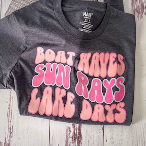 Peut inclure: Un t-shirt gris fonc&eacute; avec un motif rose et blanc qui dit "Boat Waves Sun Rays Lake Days".