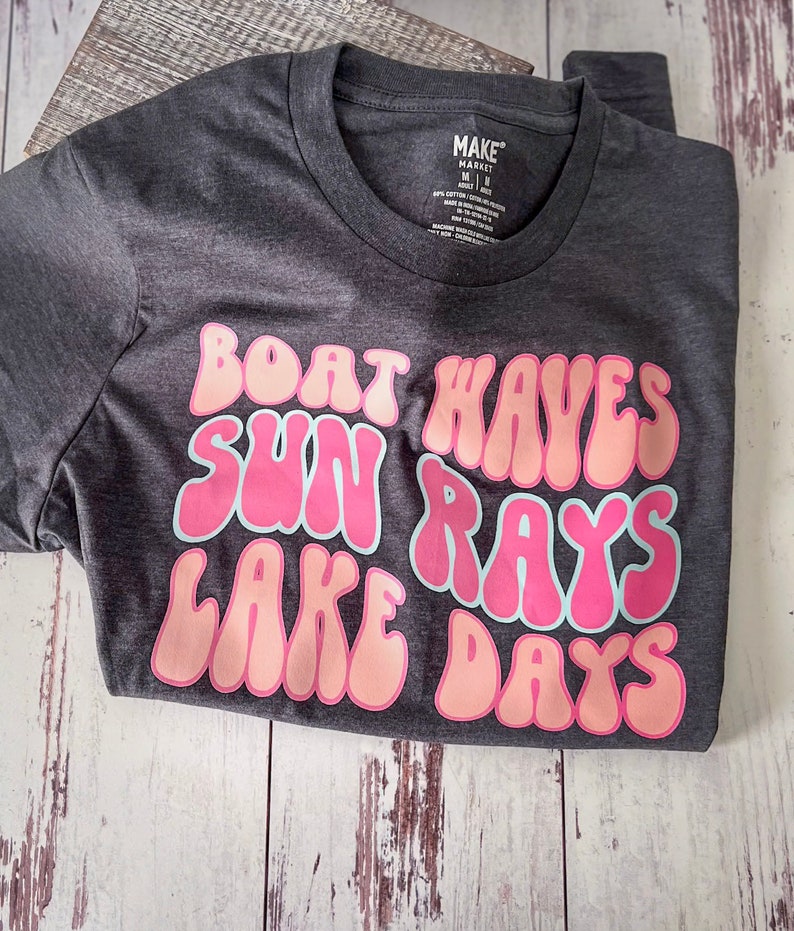 Peut inclure: Un t-shirt gris fonc&eacute; avec un motif rose et blanc qui dit "Boat Waves Sun Rays Lake Days".