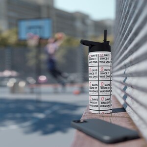 May include: A white water bottle with a black lid and straw, featuring a repeating grid pattern of black text that reads 0 DAYS SINCE IT WAS DNS, with the number 0 highlighted in red, set against a blurred outdoor basketball court background.