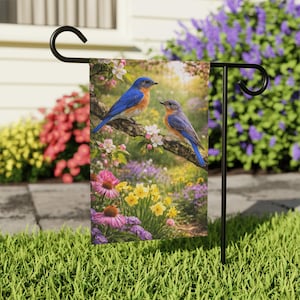 Peut inclure: Un drapeau de jardin avec une image vibrante de deux oiseaux bleus perchés sur une branche fleurie. Le drapeau présente une scène printanière colorée avec diverses fleurs roses, jaunes et violettes, sur fond de verdure luxuriante.