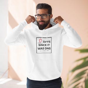 May include: A white hooded sweatshirt featuring a black square graphic on the chest. Inside the square, the text reads 0 DAYS SINCE IT WAS DNS, with the number 0 underlined in red. Below the main text, smaller text reads (It's always DNS).