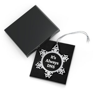 May include: A silver snowflake ornament with a black circle in the center that says "It's Always DNS". The ornament is in a black box with a silver string.
