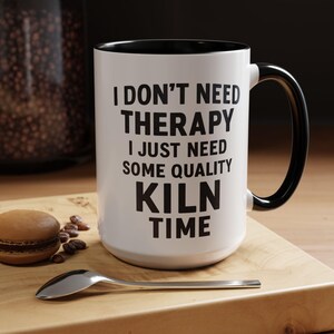 Puede incluir: Taza de cerámica blanca con interior y asa negros. La taza tiene el texto "I DON'T NEED THERAPY I JUST NEED SOME QUALITY KILN TIME" en negro. Una cuchara y un macaron están sobre una superficie de madera.