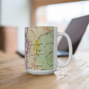May include: White ceramic mug featuring a detailed map of a mountain range, with names like Mt. Lafayette and Mt. Lincoln. The map is in shades of yellow, green, and brown. The mug has a large handle and is set on a wooden surface.