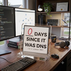 May include: A clear acrylic desk sign featuring a white square insert with black text that reads 0 DAYS SINCE IT WAS DNS (It's always DNS). The sign is positioned on a wooden desk in front of computer monitors, a keyboard, and a coffee mug.