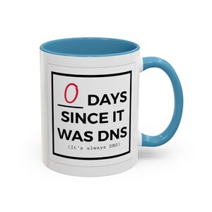 May include: White ceramic mug with a blue rim and handle. The mug features a black square with the text "0 DAYS SINCE IT WAS DNS (It's always DNS)".