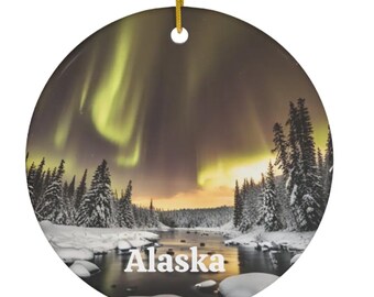 May include: Round ceramic ornament featuring a scenic view of Alaska's aurora borealis. The image shows a river, snow-covered trees, and the text "Alaska". The ornament has a gold string for hanging.