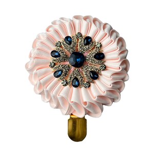 Puede incluir: Un accesorio circular con tela de satén rosa pálido plegada en un patrón de abanico. El centro está adornado con un broche de tono dorado engastado con piedras azul oscuro en forma de lágrima y cristales transparentes brillantes.