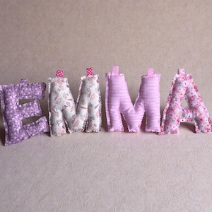Può includere: Il nome "EMMA" è scritto con lettere di stoffa. La "E" è una stampa floreale lavanda, le "M" sono a righe rosa e bianche e la "A" è una stampa floreale rosa. Le "M" hanno fiocchi di nastro a pois rosa.